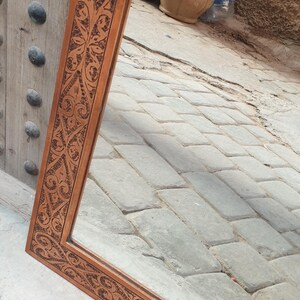 May include: A rectangular copper-colored mirror with an ornate, carved floral design around the frame. The mirror reflects a glimpse of a courtyard with stone pavers and terracotta pots.