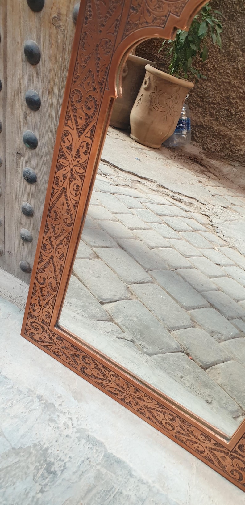 May include: A rectangular copper-colored mirror with an ornate, carved floral design around the frame. The mirror reflects a glimpse of a courtyard with stone pavers and terracotta pots.