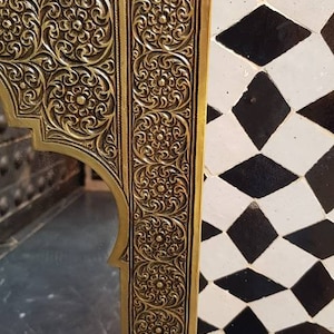 Pode incluir: Um espelho de metal dourado com uma moldura ornamentada, intrincada e esculpida. A moldura tem um design floral e est&aacute; colocada sobre um fundo de azulejos pretos e brancos.