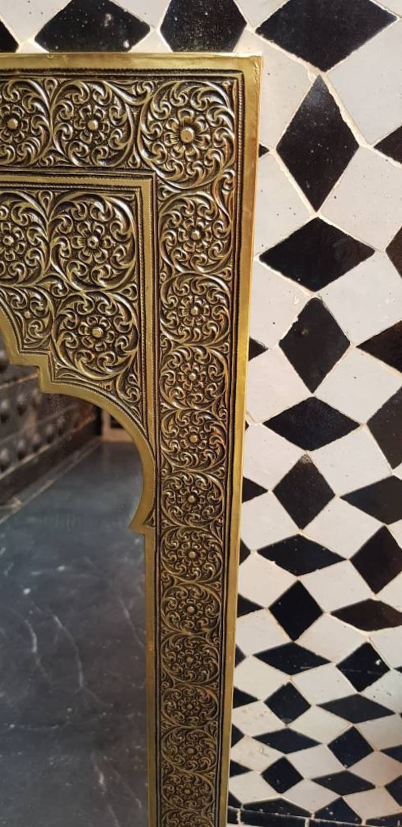 Pode incluir: Um espelho de metal dourado com uma moldura ornamentada, intrincada e esculpida. A moldura tem um design floral e est&aacute; colocada sobre um fundo de azulejos pretos e brancos.