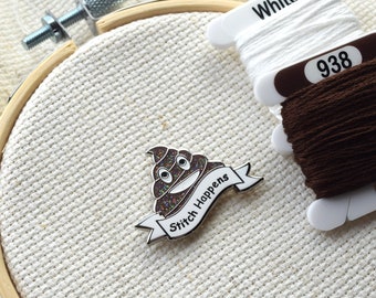 Könnte beinhalten: Eine braune und weiße Emaille-Nadel mit einem Kack-Emoji und dem Text "Stitch Happens". Die Nadel befindet sich auf einem weißen Aida-Stoff mit braunem und weißem Stickgarn.