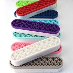 May include: A stack of colorful, rectangular ice cube trays. Each tray features a white base with a colored, patterned top in shades of green, red, blue, purple, turquoise, pink, cream, and brown. The trays are stacked on a white surface.
