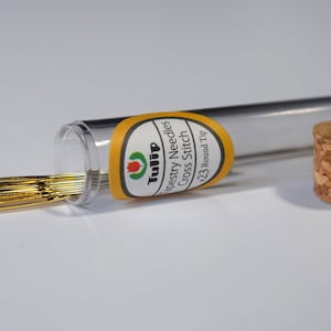 May include: A clear plastic tube with a cork lid containing a set of gold tapestry needles. The label on the tube reads "Tulip Tapestry Needles Cross Stitch #23 Round Tip".