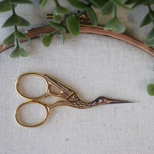 May include: Gold-toned embroidery scissors with a bird-shaped handle and a pointed tip, resting on a white fabric surface.