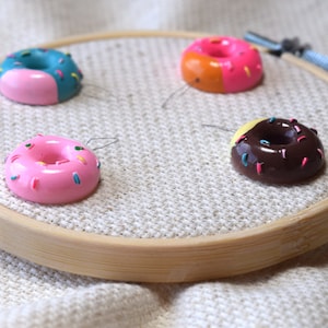May include: Four miniature donut-shaped needle threaders on a wooden embroidery hoop. The donuts are pink with colorful sprinkles, blue with pink frosting and sprinkles, orange with pink frosting and sprinkles, and brown with yellow frosting and pink sprinkles.