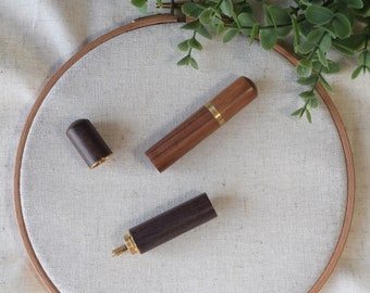 May include: Two wooden needle threaders, one with a dark brown finish and a gold tip, and the other with a lighter brown finish and a gold band. The threaders are on a white fabric background.