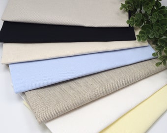 Könnte beinhalten: Ein Stapel verschiedener Aida-Stoffe in verschiedenen Farben für Kreuzstich- und Stickprojekte. Die Stoffe sind beige, schwarz, hellblau, beige, weiß und gelb.