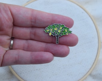 May include: A silver enamel pin featuring a green tree with yellow lemons. The pin is held in a hand.