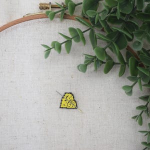 May include: A yellow and black embroidered design, possibly a patch or small artwork, is centered on a cream-colored fabric stretched within a wooden embroidery hoop. Green leafy branches frame the top right corner.