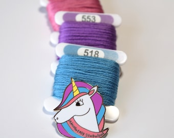 May include: A gold-tone enamel pin featuring a cartoon unicorn with a pink, blue, and purple mane and a yellow horn. The unicorn is wearing a banner that reads "Absolutely Stabulous".