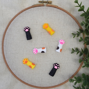 May include: A collection of colorful cat paw-shaped charms. The charms are in various colors, including black, yellow, and white, with pink and orange paw pads. They are arranged on a beige fabric stretched within a wooden embroidery hoop.