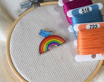 Könnte beinhalten: Ein blauer Vogel sitzt auf einem Regenbogen vor weißem Hintergrund. Der Regenbogen besteht aus sechs Farben: Rot, Orange, Gelb, Grün, Blau und Lila. Der Vogel schaut nach rechts. Das Bild ist auf weißem Hintergrund.
