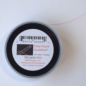 May include: A spool of red thread with the text "Easy-Count Guideline" and "100 yards RED". The spool is black with a white label and a thin strand of red thread extending from it.