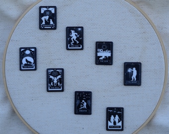 Könnte beinhalten: Ein Satz von zehn schwarzen und weißen Tarotkarten mit Illustrationen von Figuren und Symbolen. Die Karten sind in einem Kreis auf einem weißen Stoffhintergrund angeordnet.