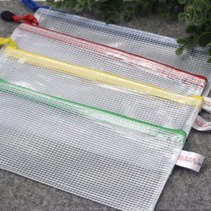 May include: Four clear mesh zipper pouches with colored zippers in green, yellow, red, and blue. Each pouch has a matching colored zipper pull and a small white tag. The pouches are arranged diagonally on a gray surface.