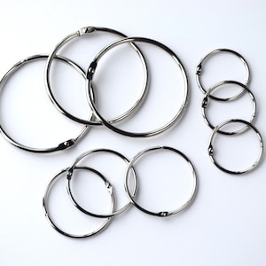 May include: A collection of silver metal binder rings of varying sizes, arranged on a white surface. The rings are open, showing their hinged design. The image showcases the rings' shiny, metallic appearance and their potential for organizing documents or other items.