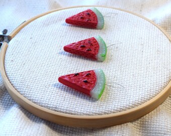 Könnte beinhalten: Drei rote und grüne Wassermelonenstücke mit schwarzen Kernen sind auf einem weißen Stoffhintergrund in einem hölzernen Stickrahmen befestigt.