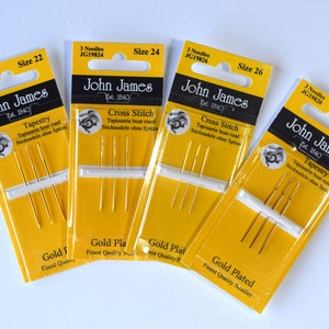 May include: Four packages of gold-plated needles for tapestry and cross stitch. The packages are yellow and feature a black and white illustration of a dragon. The packages are labeled with the brand name "John James" and the size of the needles. The sizes are 20, 22, 24, and 26.