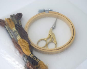 May include: A white embroidery hoop with a wooden frame, a pair of gold embroidery scissors, and a variety of embroidery floss in brown, yellow, and gray colors.