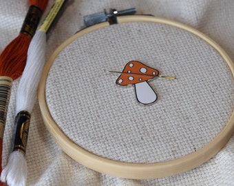Könnte beinhalten: Nahaufnahme eines weißen Stickrahmens mit einer roten und weißen Pilznadel darin. Der Rahmen liegt auf einer weißen Stoffoberfläche. Links vom Rahmen befinden sich zwei Spulen mit Stickgarn, eine orange und eine weiße.