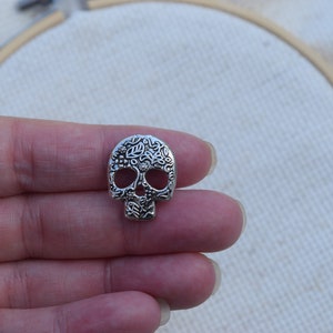 Könnte beinhalten: Silberner Totenkopf-Anhänger mit floralem Design. Der Totenkopf ist mit filigranen Schnitzereien verziert und hat eine traditionelle Zucker-Schädel-Ästhetik.