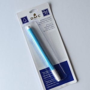 May include: A D.M.C. Embroidery Transfer Pen in a clear plastic package. The pen is light blue with a white tip. The packaging includes text in English and French, describing the pen's features, including water-soluble ink.