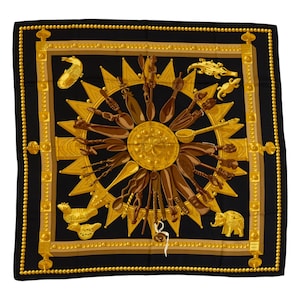 Könnte beinhalten: Ein quadratisches Seidentuch mit schwarzem Hintergrund und einem zentralen goldenen Sonnenmotiv, umgeben von verschiedenen braunen Holzutensilien und Werkzeugen. Der Rand ist mit goldenen Perlenlinien und geometrischen Mustern verziert.