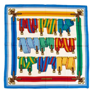 Puede incluir: Pañuelo cuadrado de seda con fondo crema y borde azul. El pañuelo presenta filas de cintas de colores rojo, azul, verde y amarillo, colgadas de barras doradas. El texto "HERMES-PARIS" es visible.