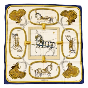 Puede incluir: Pañuelo cuadrado de seda con fondo crema y borde azul marino. El pañuelo presenta una imagen central enmarcada de un carruaje tirado por caballos, rodeada de ilustraciones de caballos y diseños de silla de montar dorados. El texto "GRAND APPARAT" es visible.