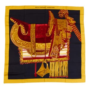 Puede incluir: Pañuelo cuadrado de seda con diseño de silla de montar marroquí. El pañuelo presenta un fondo negro con una silla de montar roja y dorada, flecos dorados y un borde amarillo. El texto "SELLE D'APPARAT MAROCAINE" está en la parte superior.
