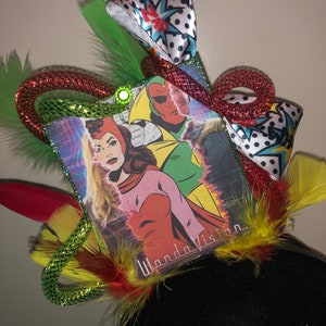May include: A black headband with a red, green, and yellow feather trim. A square card with a comic book image of Wanda and Vision is attached to the headband. The card has the text "Wanda Vision" on it.