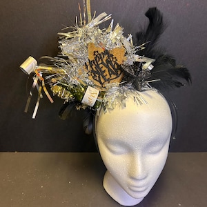 May include: A festive headpiece featuring a champagne bottle, silver tinsel, black feathers, and a gold star with "Happy New Year" text. The headpiece is mounted on a white mannequin head.