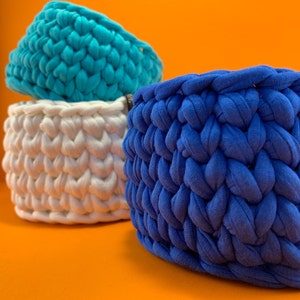 Può includere: Tre cesti in crochet di diversi colori: turchese, bianco e blu. I cesti sono realizzati con filato spesso e hanno un motivo intrecciato testurizzato.
