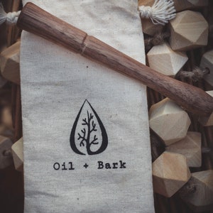 May include: A small, white canvas bag with a wooden stirring stick inside. The bag has a black tree logo and the text "Oil + Bark".