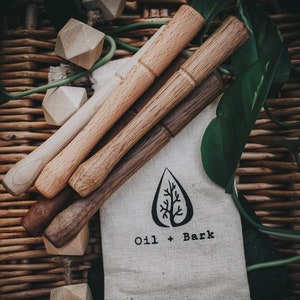 May include: A set of four wooden stirring sticks in a white fabric bag with the text "Oil + Bark". The sticks are made of different types of wood, including light and dark wood.