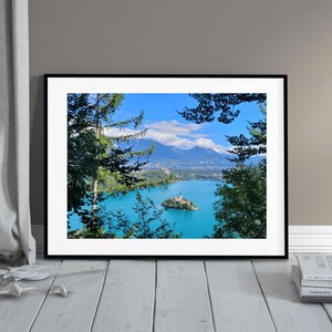 Könnte beinhalten: Ein gerahmtes Foto eines Sees mit einer kleinen Insel in der Mitte. Auf der Insel steht eine Kirche. Der See ist von Bäumen und Bergen in der Ferne umgeben. Der Himmel ist blau mit weißen Wolken.