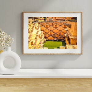 May include: An aerial view of a large, intricately designed stepwell in India. The stepwell is made of light brown stone and has a central pool of water. The steps lead down to the water, and there are small alcoves and niches along the sides.