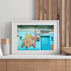 May include: Framed artwork featuring a vibrant mural of an elephant carrying figures, painted on a light blue wall. The elephant is adorned with intricate patterns and colors. A red flag and a turquoise door are also visible.