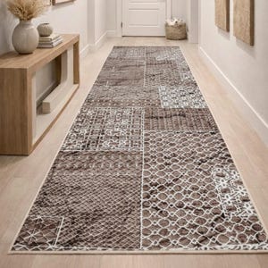 Op de afbeelding: Een lange, rechthoekige bruin-wit patroonkleed in een gang. Het kleed heeft een patchwork-ontwerp met verschillende geometrische en getextureerde patronen. De gang heeft lichtgekleurde muren, een houten console en kunstwerken.