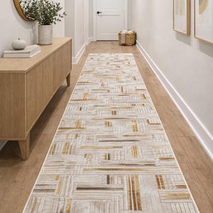 May include: A rectangular hallway rug with a geometric pattern in shades of beige, brown, and gold. The rug is placed on a wooden floor in a hallway. The rug's design features a series of intersecting lines. The hallway has a wooden cabinet and a mirror.