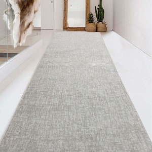 May include: A long, rectangular area rug featuring a light grey, textured, woven pattern. The rug is positioned centrally on a smooth, white floor, extending from the foreground towards the back of the room, creating a clean, neutral visual path.