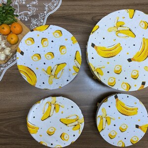 May include: Four round, white fabric food covers with a yellow banana print. The covers are different sizes and are arranged on a wooden surface.