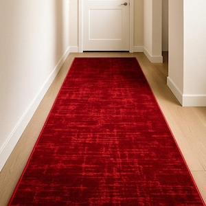 May include: A long, rectangular red rug with a textured pattern, placed in a hallway with light-colored walls and a wooden floor. The rug's deep red color provides a striking contrast to the neutral tones of the space.