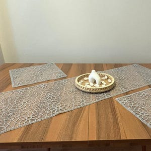 Pode incluir: Uma mesa de madeira com um caminho de mesa de renda bege e dois jogos americanos a condizer. Uma bandeja decorativa com uma escultura branca e pequenas pedras está sobre o caminho.