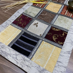 Puede incluir: Un camino de mesa rectangular con un diseño de patchwork. El camino de mesa está hecho de diferentes telas con varios patrones y colores, incluyendo beige, marrón, rojo y negro. El camino de mesa tiene un borde blanco con un patrón texturizado.