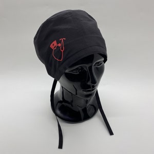 Peut inclure: Un bonnet de chirurgien noir avec un symbole médical rouge et un stéthoscope sur le devant. Le bonnet a des liens noirs.
