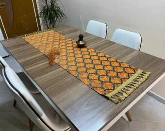Puede incluir: Una mesa de madera marrón con un mantel blanco con flecos dorados. El mantel tiene un patrón geométrico en naranja y negro. Hay cuatro sillas blancas alrededor de la mesa.