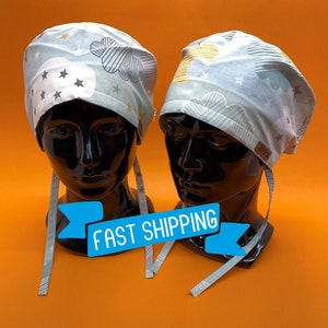 Puede incluir: Dos gorros quirúrgicos de color azul claro con un patrón de nubes y estrellas blancas y grises. Los gorros tienen lazos largos y se muestran en cabezas de maniquí negras. El texto "ENVÍO RÁPIDO" se muestra en una pancarta azul.