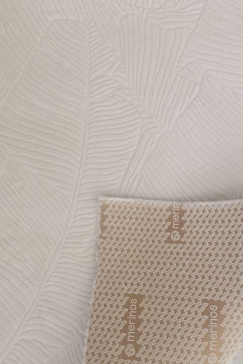 May include: A white rug with a textured leaf pattern. The rug has a brown backing with the word "merinos" printed on it.