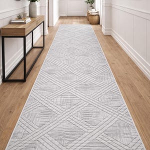 Op de afbeelding: Een lange, rechthoekige grijze loper met een geometrisch patroon van elkaar kruisende lijnen. De loper ligt in een gang met een lichte houten vloer en witte muren. Het ontwerp van de loper voegt een moderne touch toe aan de ruimte.